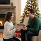 Man and woman exchanging trading card deck box gifts in a cozy living room with a Christmas tree and fireplace.